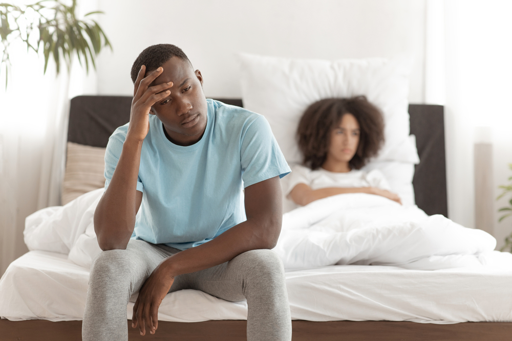 Sad frustrated young african american guy sits on bed, offended female looks to side in bright interior of bedroom.