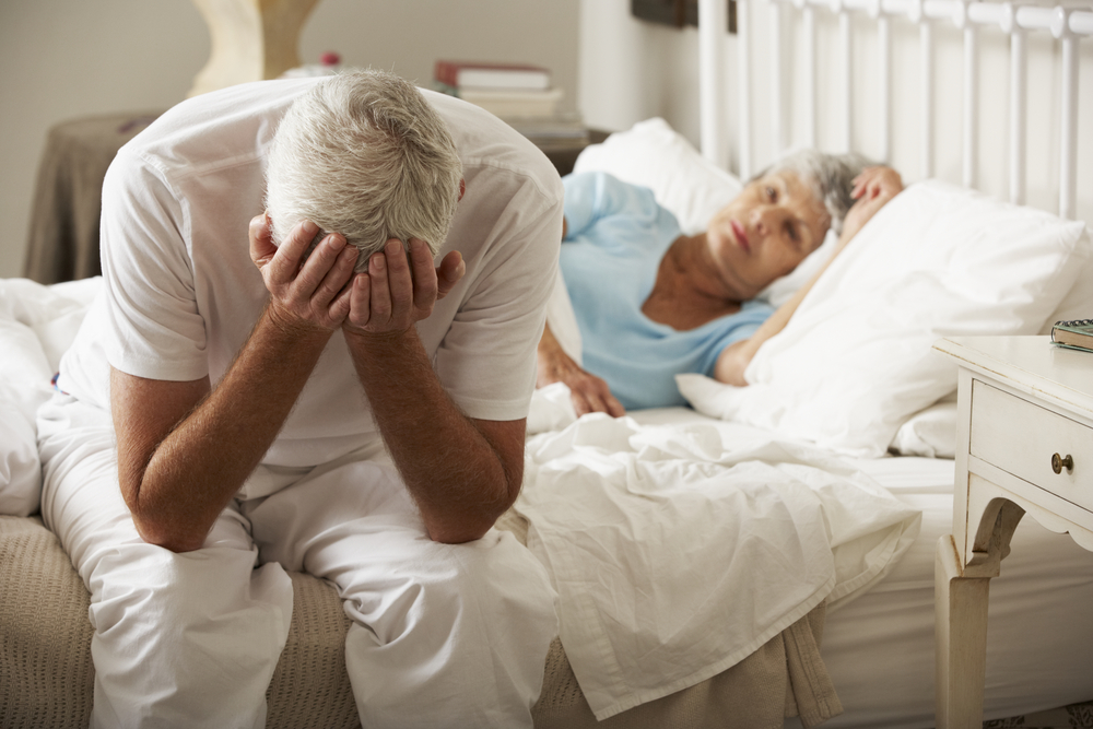 Worried Senior Man Sits On Bed Next To Wife.